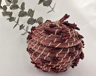 May include: A small, round, lidded basket woven from reddish-brown and cream-coloured fibres. The basket has a textured surface with a decorative, fringed edge. Dried eucalyptus leaves are in the background.