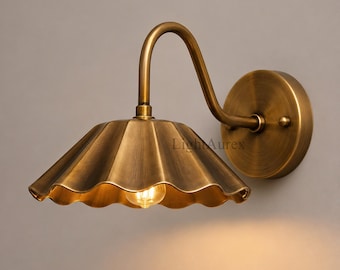 May include: A wall-mounted lamp featuring a curved arm and a scalloped, cone-shaped shade in a brushed brass finish. The fixture includes a circular wall plate and a single exposed light bulb emitting a warm glow. The text LightAurex is visible.