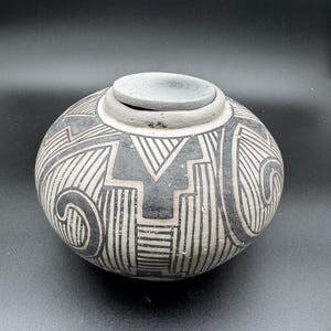 Op de afbeelding: Een ronde, met de hand gevormde keramische pot met bijpassend deksel, voorzien van een matte afwerking in grijze, zwarte en witte tinten. De buitenkant is versierd met complexe, handgeschilderde geometrische patronen, waaronder scherpe zigzaglijnen en bochten.