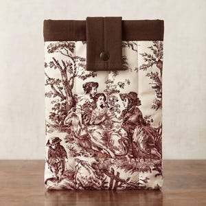 May include: A rectangular quilted fabric sleeve featuring a brown and cream toile de jouy pattern depicting figures in a landscape. The sleeve has a solid brown fabric top border and a central brown fabric tab closure secured with a matching snap button.