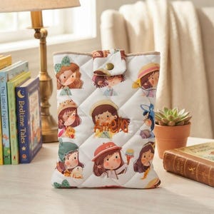 May include: A white quilted fabric pouch featuring a repeating pattern of illustrated characters wearing various hats and accessories. The center of the pouch displays the embroidered name Amella in orange thread and includes a snap button closure.