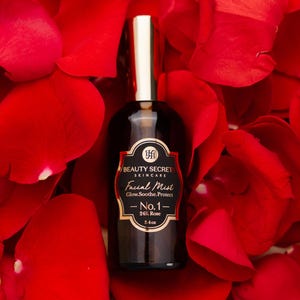 Puede incluir: Un frasco de vidrio oscuro de bruma facial Beauty Secrets Skincare No.1 24K Rose con tapa dorada, etiquetado 3.4oz, rodeado de p&eacute;talos de rosa rojos. El texto dice: Complete Your Routine y Pair with our 24K Rose Treatment Mist.