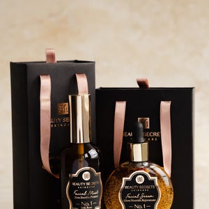 May include: A dark amber facial mist bottle and a round facial serum bottle with gold flakes, both labeled Beauty Secrets Skincare 24K Rose No. 1. The mist is 3.4 oz and the serum is 1.7 oz, presented with two black gift boxes featuring pink ribbon handles.