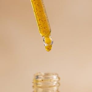 Puede incluir: Un cuentagotas de vidrio transparente lleno de un l&iacute;quido amarillo dorado que contiene peque&ntilde;as escamas doradas brillantes. Una gota de la sustancia viscosa y brillante cuelga de la punta del cuentagotas, sobre el cuello abierto de una botella.