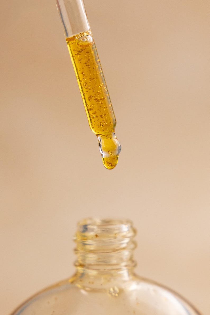 Puede incluir: Un cuentagotas de vidrio transparente lleno de un l&iacute;quido amarillo dorado que contiene peque&ntilde;as escamas doradas brillantes. Una gota de la sustancia viscosa y brillante cuelga de la punta del cuentagotas, sobre el cuello abierto de una botella.