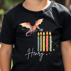 Könnte beinhalten: Ein Kind trägt ein schwarzes T-Shirt mit einem bunten Drachenmotiv und dem Namen "Henry" in Schreibschrift. Das Shirt zeigt auch Geburtstagskerzen. Im Hintergrund sind grüne und orangefarbene Ballons zu sehen.