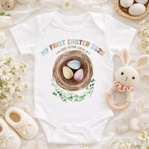 Puede incluir: Body de bebé blanco con el texto "MY FIRST EASTER 2026" y un diseño de nido de acuarela con tres huevos. Un sonajero de conejito de ganchillo y zapatos de bebé están al lado del body. Ropa de bebé con temática de Pascua.