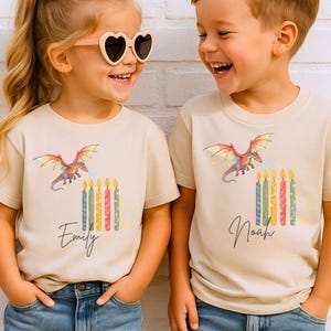 Puede incluir: Dos niños con camisetas beige a juego con un diseño de dragón y velas. Las camisetas tienen los nombres "Emily" y "Noah" impresos. La niña lleva gafas de sol en forma de corazón y un lazo.
