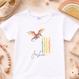 Puede incluir: Camiseta blanca con un dragón colorido soplando fuego a velas de cumpleaños. El nombre "Joshua" está escrito en cursiva. La camiseta está rodeada de juguetes y objetos decorativos.