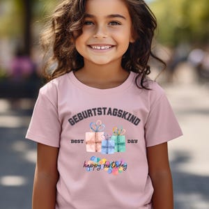 May include: A young girl wearing a light pink t-shirt with the words "GEBURTSTAGSKIND" and "happy birthday" printed on it. The shirt features watercolor illustrations of wrapped gifts in pastel colors. The girl is smiling.