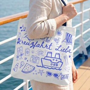 Può includere: Una borsa tote bianca con illustrazioni e testo blu, tra cui una nave da crociera, occhiali da sole e le parole "Kreuzfahrt Liebe". La borsa è portata da una persona sul ponte di una nave da crociera, con il mare sullo sfondo.
