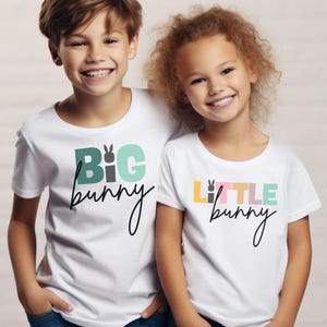May include: Two children wearing white t-shirts with Easter-themed designs. One shirt says "BIG bunny" in green, gray, and light blue, and the other says "LITTLE bunny" in yellow, pink, and light green. Both shirts have a bunny graphic.