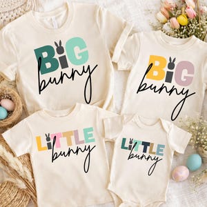 May include: Four cream-colored t-shirts and a onesie with Easter-themed text. The shirts and onesie feature the words "BIG bunny" and "LITTLE bunny" in colorful block letters and a bunny graphic. The text is in black script.