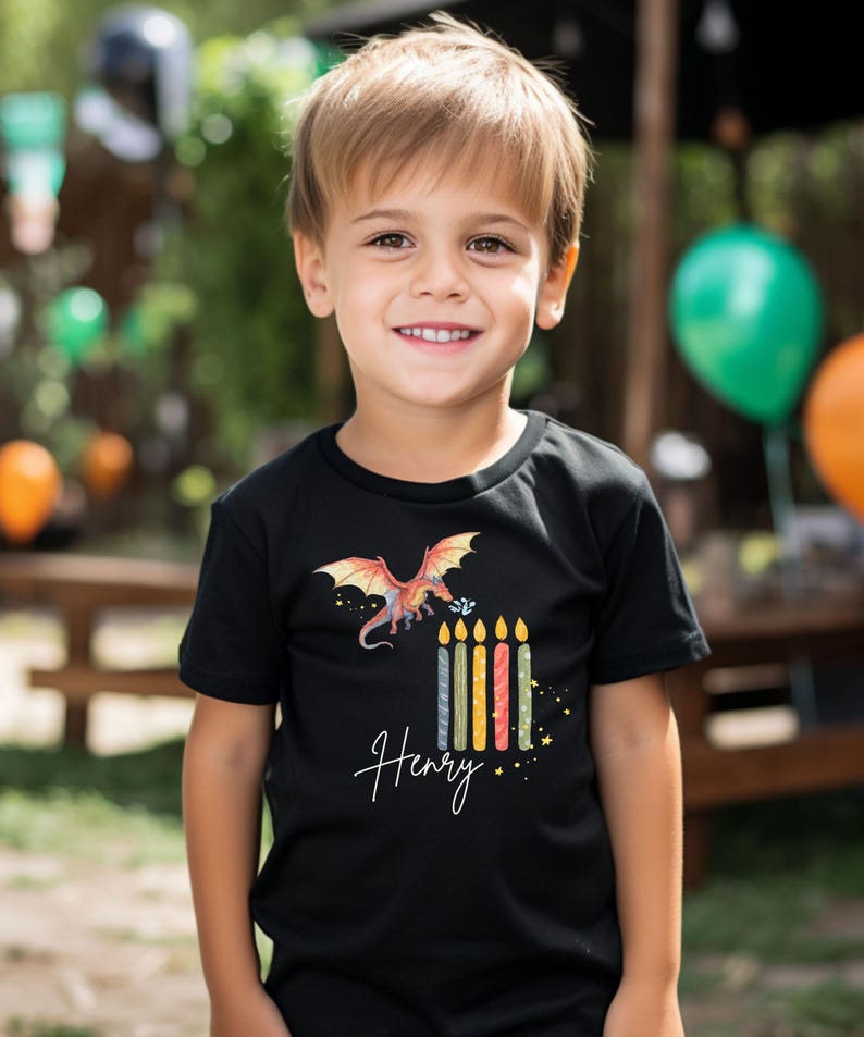 Könnte beinhalten: Ein Kind trägt ein schwarzes T-Shirt mit einem bunten Drachenmotiv und dem Namen "Henry" in Schreibschrift. Das Shirt zeigt auch Geburtstagskerzen. Im Hintergrund sind grüne und orangefarbene Ballons zu sehen.