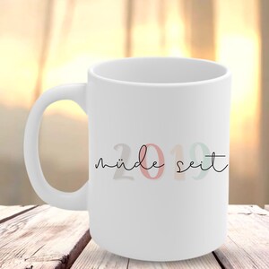 May include: White ceramic mug with a curved handle. The mug features the text "müde seit" in black script, with the numbers "2019" in pastel colours. The mug is set on a wooden surface.