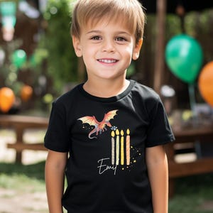 May include: A black short-sleeved t-shirt featuring a graphic of a red and orange dragon flying above four lit birthday candles. The name "Emil" is printed in white cursive script below the candles, surrounded by small yellow star accents.
