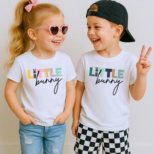 May include: Two children wearing white t-shirts with the text "Little Bunny" in colorful letters. One child wears heart-shaped sunglasses and a pink bow. The other child wears a black baseball cap and checkered shorts.
