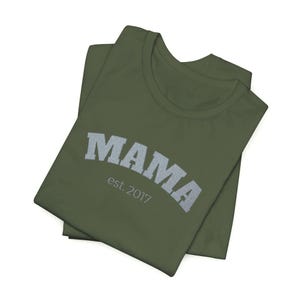 Puede incluir: Camiseta verde oliva con la palabra "MAMA" en gris, en forma de arco. Debajo de la palabra "MAMA" está el texto "est. 2017" en una fuente gris más pequeña. La camiseta está doblada.