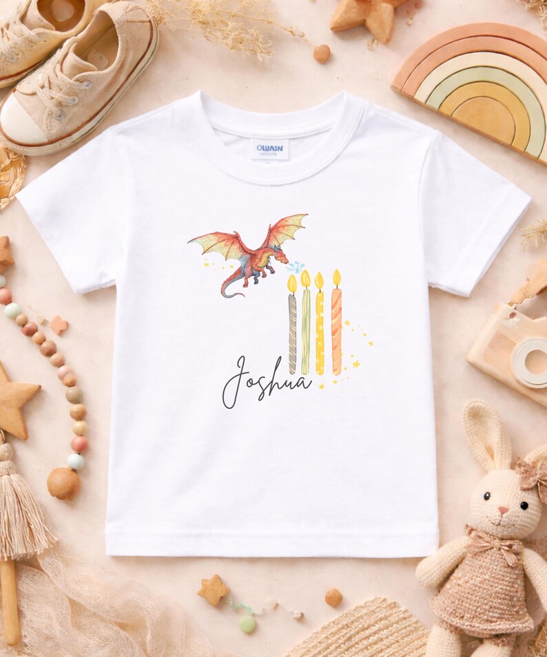 Könnte beinhalten: Weißes T-Shirt mit einem bunten Drachen, der Feuer auf Geburtstagskerzen speit. Der Name "Joshua" ist in Schreibschrift geschrieben. Das T-Shirt ist von Spielzeug und dekorativen Gegenständen umgeben.