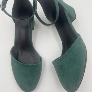 Op de afbeelding: Een paar smaragdgroene su&egrave;de enkellaarsjes met enkelbandjes. De schoenen hebben een ronde neus, een kleine blokhak en een gespsluiting op het enkelbandje. De binnenkant van de schoen is zwart.