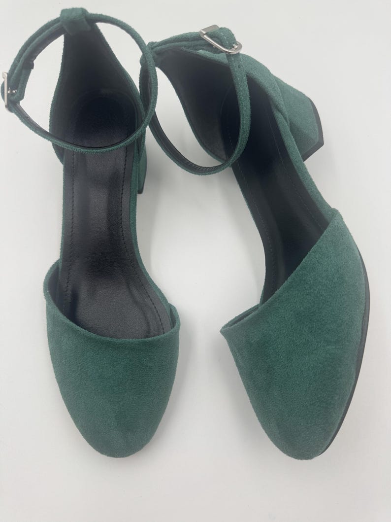 Op de afbeelding: Een paar smaragdgroene su&egrave;de enkellaarsjes met enkelbandjes. De schoenen hebben een ronde neus, een kleine blokhak en een gespsluiting op het enkelbandje. De binnenkant van de schoen is zwart.