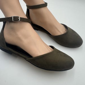 May include: Olive green flats with ankle straps. These shoes feature a rounded toe and a small buckle closure on the ankle strap. The flats are made of a soft, suede-like material and have a simple, elegant design.