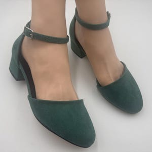 Op de afbeelding: Een paar donkergroene su&egrave;de enkellaarzen met enkelband. De schoenen hebben een ronde neus, een lage blokhak en een verstelbare enkelband met een zilveren gesp. Op een witte ondergrond.