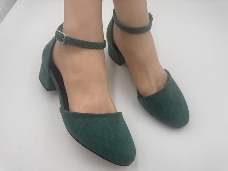 Op de afbeelding: Een paar donkergroene su&egrave;de enkellaarzen met enkelband. De schoenen hebben een ronde neus, een lage blokhak en een verstelbare enkelband met een zilveren gesp. Op een witte ondergrond.