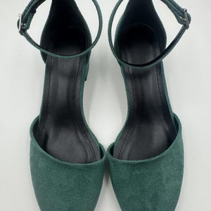 Op de afbeelding: Een paar groene su&egrave;de enkelsandalen. De open teen schoenen hebben een dunne enkelband met een zilveren gesp. De binnenkant van de schoen is zwart. De sandalen worden op een witte achtergrond gepresenteerd.