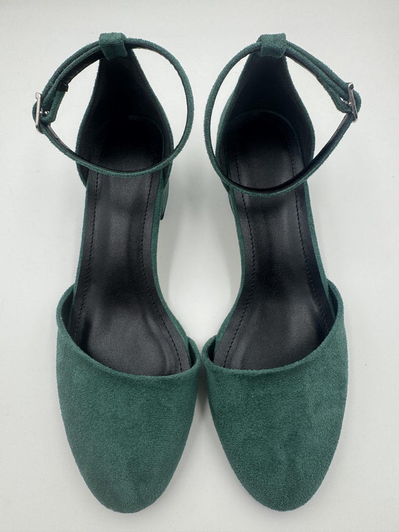 Op de afbeelding: Een paar groene su&egrave;de enkelsandalen. De open teen schoenen hebben een dunne enkelband met een zilveren gesp. De binnenkant van de schoen is zwart. De sandalen worden op een witte achtergrond gepresenteerd.