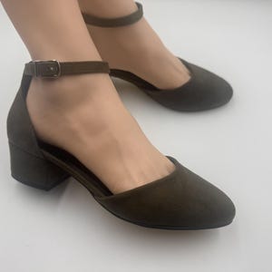 May include: Olive green ankle strap heels with a block heel. The open-sided shoes have a rounded toe and a buckle closure on the ankle strap. The heels are approximately 2 inches high.