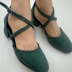 May include: A pair of dark green suede sandals with a low block heel. The sandals feature a crisscross strap design across the foot and an ankle strap with a silver buckle. The shoes are displayed on a white background.