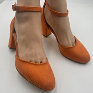 Könnte beinhalten: Ein Paar orangefarbene Wildleder-Sandalen mit Absatz. Die Schuhe haben eine runde Spitze, einen Knöchelriemen und einen Blockabsatz. Die Schuhe werden auf einer weißen Oberfläche präsentiert.