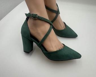 May include: Emerald green suede heels with a pointed toe and a thick block heel. The shoes feature a crisscross ankle strap with a gold-tone buckle. The heels are approximately 3 inches high.