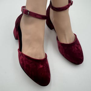 Könnte beinhalten: Ein Paar bordeauxfarbene Samt-Knöchelriemenschuhe mit niedrigem Blockabsatz. Die Schuhe haben eine runde Spitze und einen Schnallenverschluss am Knöchelriemen. Die Schuhe werden auf einer weißen Oberfläche präsentiert.
