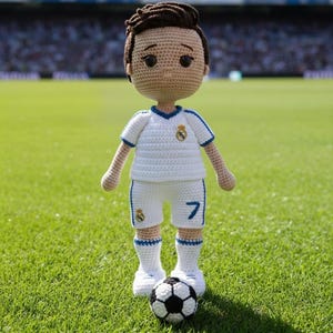 Puede incluir: Muñeco de ganchillo de un futbolista con uniforme blanco y detalles azules, con el número 7. Está de pie en un campo verde con un balón de fútbol. Se ve un estadio al fondo.