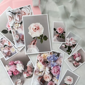 May include: A collection of floral art prints featuring roses and other flowers in shades of pink, white, and blue. The prints are arranged with pink envelopes and a light gray ribbon on a white surface. The images showcase detailed floral arrangements.
