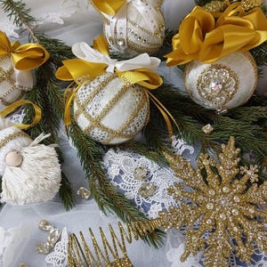 May include: A festive arrangement of Christmas ornaments and decorations. The image features white and gold ornaments with intricate detailing, gold bows, and a large gold snowflake. A small doll and pinecone are also present.