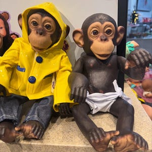May include: Two chimpanzee dolls. One wears a yellow raincoat with blue buttons and denim jeans. The other chimpanzee doll is dark brown and wears a white diaper. Both dolls have realistic facial features and are sitting side by side.