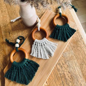May include: Three macrame ornaments featuring smooth wooden rings and natural wooden beads. Two ornaments are crafted with dark forest green cotton cord, and one is crafted with light gray cotton cord, each ending in a fringed tassel design.