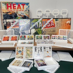 May include: A collection of board game components, including boxes labeled "HEAT: Pedal to the Metal" and expansion packs. Various game cards, trays, and game boards are arranged on a wooden surface, with a green surface in the foreground.