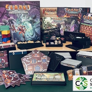 May include: A collection of board game components, including boxes, cards, and game boards. The game boxes feature the title "Clank! Catacombs" and other expansions. The components are arranged on a wooden surface with a green background.