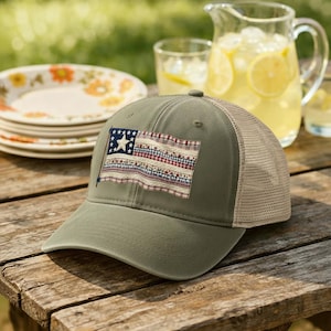 May include: An olive green trucker hat with a tan mesh back. The hat features a patch of a vintage American flag with a blue star field and red and white stripes. The hat is sitting on a wooden table.