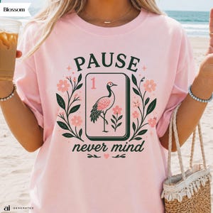 May include: A light pink t-shirt featuring a graphic of a rectangular tile with the number 1 and a crane illustration. The design includes the text PAUSE and never mind, surrounded by dark green leafy branches and small pink flowers.