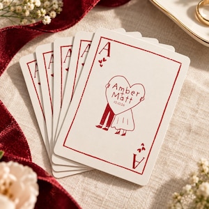 May include: A fan of white playing cards with red ink details. The top card features an Ace of Hearts design with a central heart graphic containing the text Amber + Matt and the date 10.10.26, held by two illustrated figures.