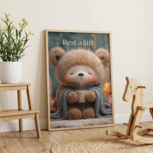 Könnte beinhalten: Gerahmter Druck mit einem Cartoon-Bären, der einen Hut und eine Decke trägt und eine Tasse hält. Der Text "Rest a little" steht oben. Der Bär ist braun mit rosigen Wangen, und die Decke ist blau.
