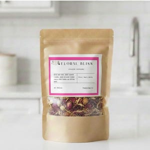Peut inclure: Un sac en papier brun de Floral Bliss, avec une étiquette rose et blanche. L'étiquette énumère des ingrédients comme la rose, le jasmin et la vanille. Le sac est partiellement transparent, révélant des fleurs et des herbes séchées.