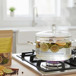 May include: A glass saucepan with a lid, containing water, citrus slices, and spices, steams on a gas hob. A packet labelled "Energy Boost" and dried citrus slices are on the counter. The kitchen has white cabinets and a window.