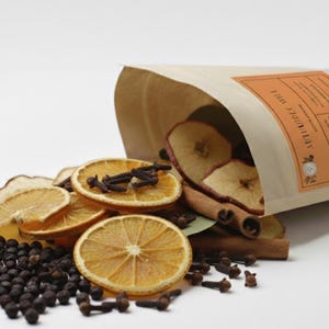 May include: A brown paper bag spills dried orange slices, apple slices, cinnamon sticks, cloves, and peppercorns. The bag has a label with text. This assortment is likely for potpourri or festive decoration.