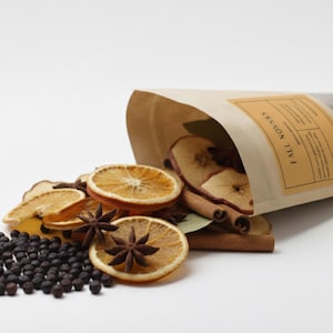 Puede incluir: Una bolsa de papel marrón con la etiqueta "FALL NONNAS" derrama rodajas de naranja secas, rodajas de manzana, anís estrellado, palitos de canela, hojas de laurel y granos de pimienta negra. Los ingredientes están dispuestos sobre una superficie blanca.